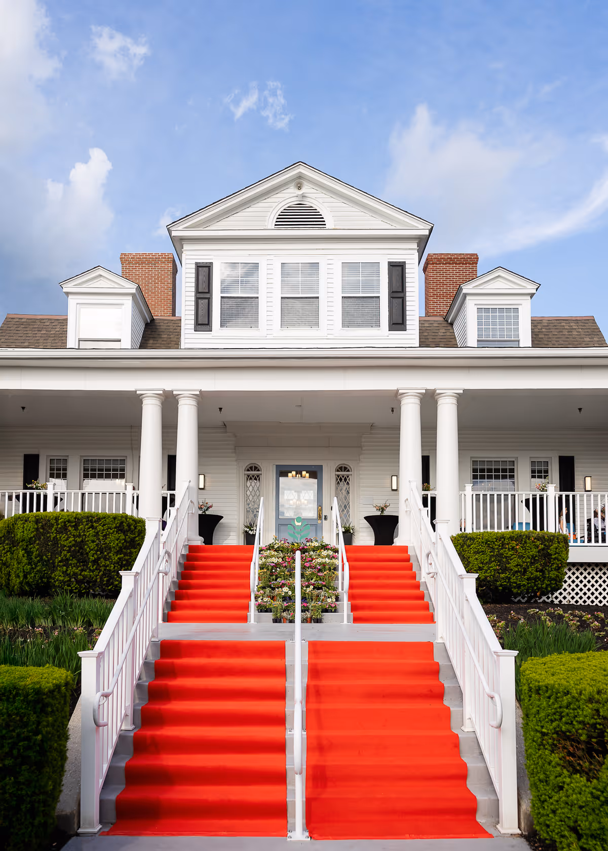 Front exterior view of a large white building with a red carpet covering the stairs leading up to the entrance. The building has white columns, multiple windows with black shutters, and neatly trimmed green bushes on either side of the stairs under a partly cloudy blue sky.