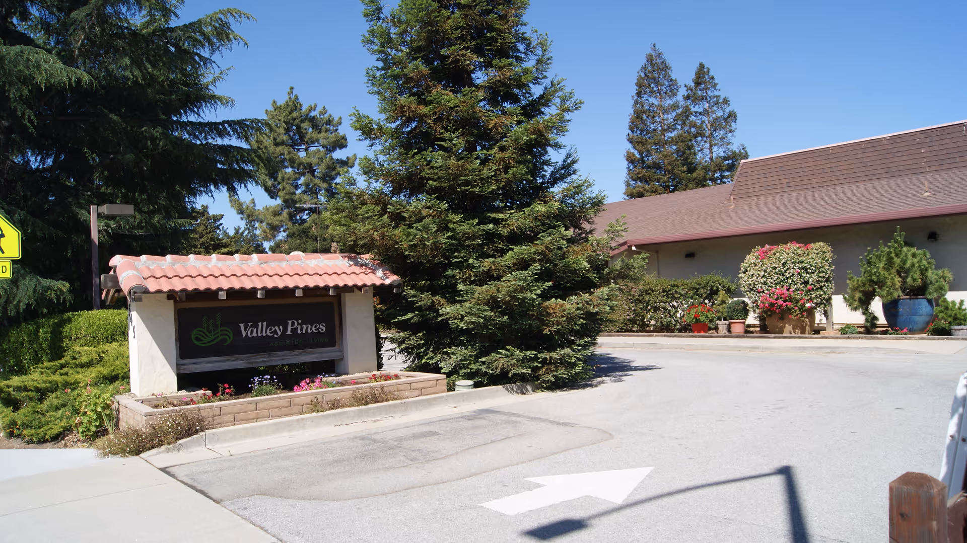 Exterior view of Valley Pines Assisted Living facility entrance with a sign displaying the facility name surrounded by greenery and trees under a clear blue sky.