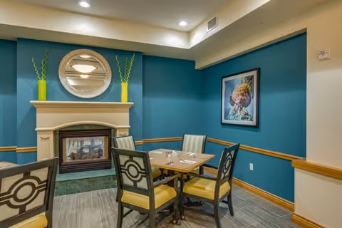 A cozy dining area with a wooden table set for four, surrounded by four chairs with yellow cushions and decorative backs. The room features teal walls with wooden trim, a fireplace with a round mirror above it, two tall green vases on the mantel, and a framed artwork on the wall.