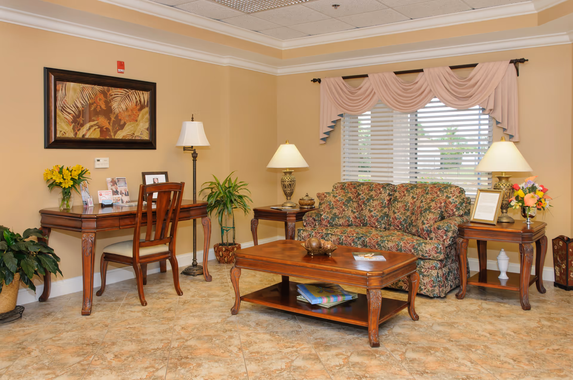 Well-lit sitting area featuring a floral upholstered sofa, wooden coffee table and matching side tables with lamps, a desk and decorative plants against beige walls.