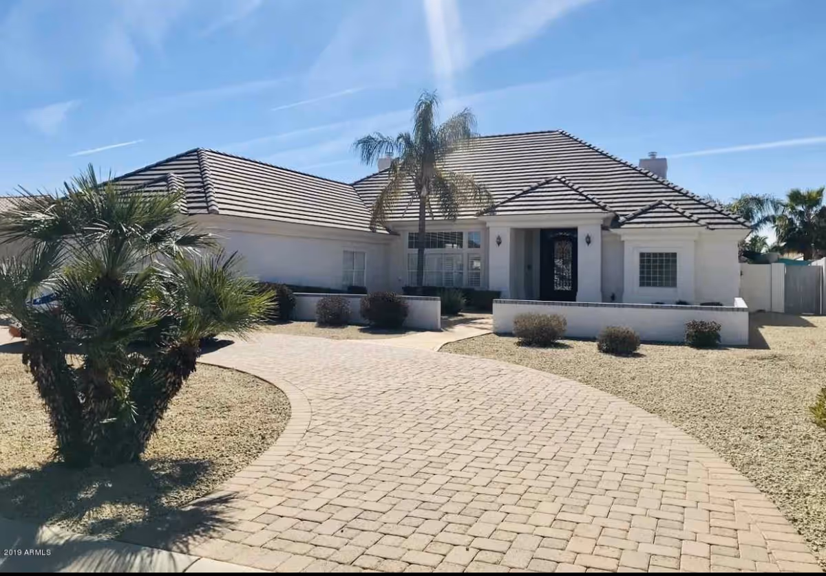 Front exterior view of a single-story white house with a tiled roof, a curved paved driveway, palm trees, and desert landscaping under a clear blue sky.