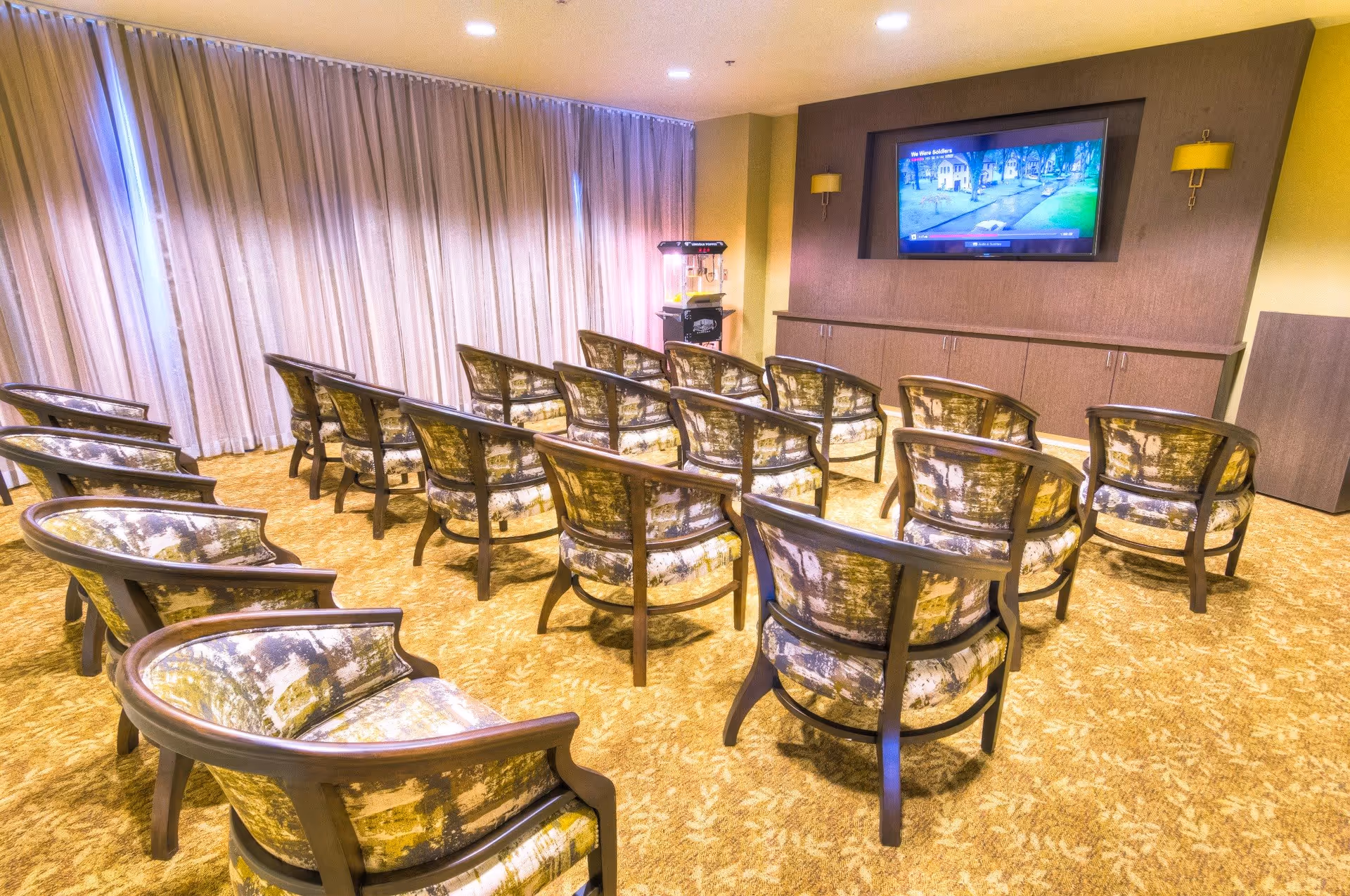 A small theater-style room with multiple rows of patterned armchairs facing a wall-mounted flat screen TV. The room has beige carpet, light-colored curtains covering the windows, and a popcorn machine in the corner.