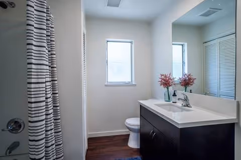 A clean bathroom featuring a shower with a striped curtain on the left, a white toilet in front of a window with frosted glass, and a dark wood vanity with a white countertop and a sink. A large mirror is mounted above the vanity, reflecting part of the room.