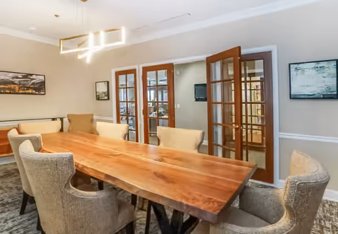 A dining room with a long wooden table surrounded by six upholstered chairs. The room features modern rectangular pendant lighting above the table, light-colored walls with white trim, and two framed artworks on the walls. There are glass-paneled double doors leading to another room.