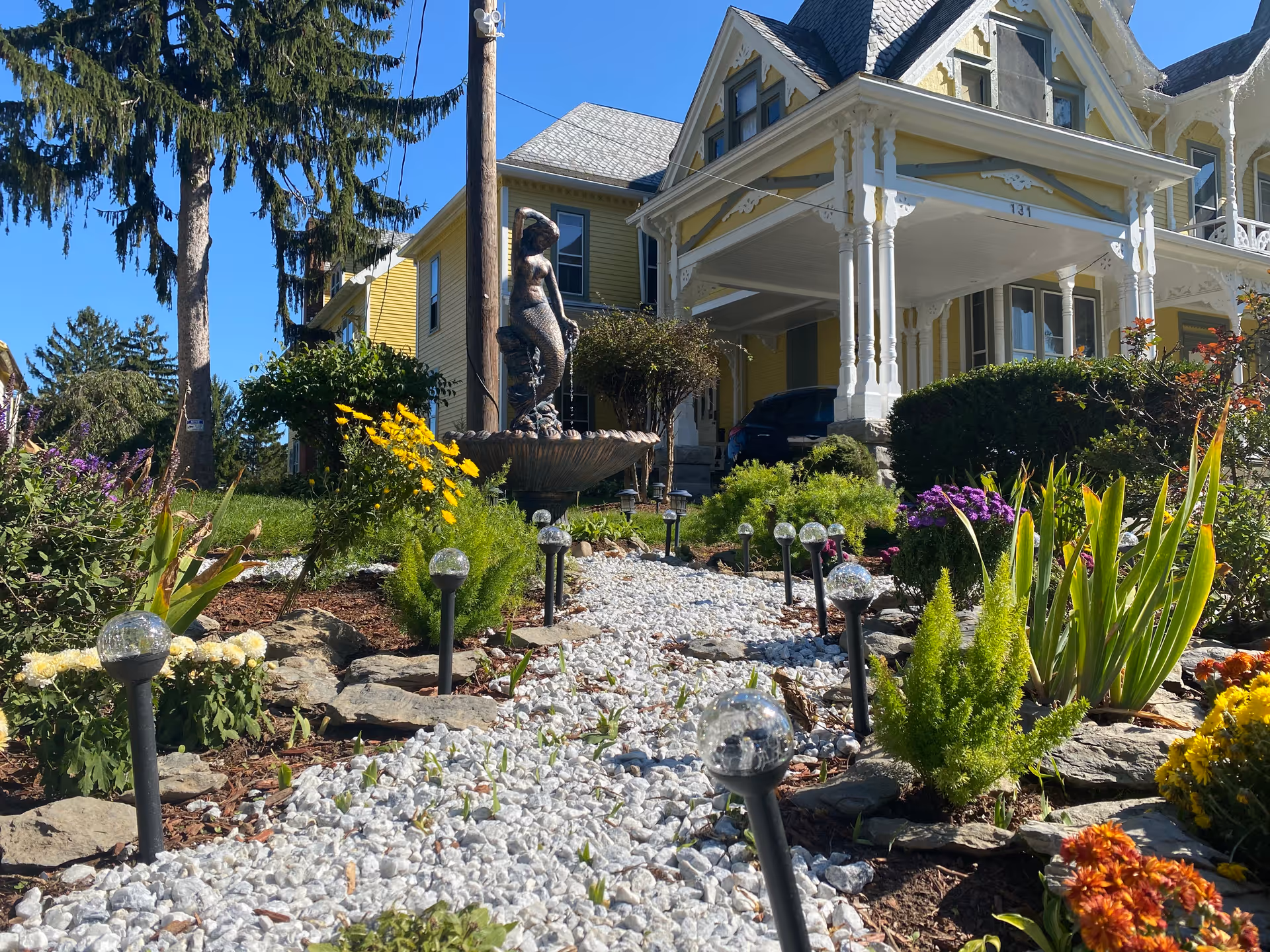 Gravel garden path lined with solar lights, flowers, a fountain statue, and a yellow Victorian house with a covered porch in the background.