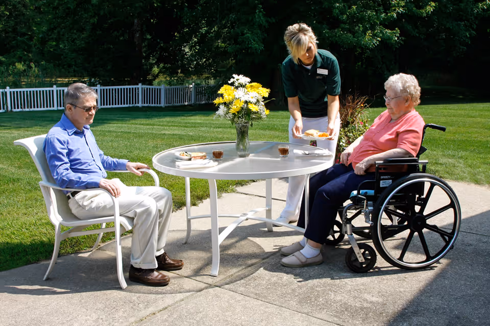An elderly man sitting on a white chair and an elderly woman in a wheelchair are seated outside at a round table with a vase of yellow and white flowers. A caregiver is serving food to the woman in the wheelchair. The setting is a sunny patio with green grass and trees in the background.