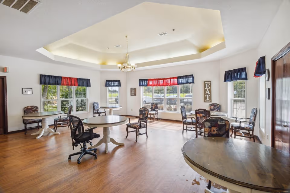 A bright dining room with multiple round tables and chairs arranged around the room. The room has large windows with red and blue valances, wooden flooring, and a chandelier hanging from a recessed ceiling. There is a sign on the wall that says 'EAT'.