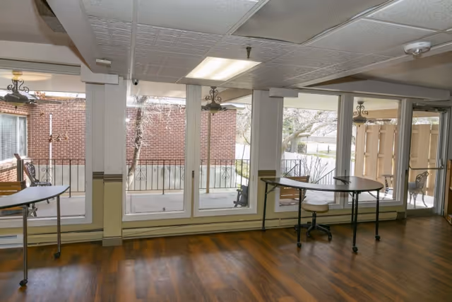 Interior room with large windows showing an outdoor patio area with chairs. Inside, there are two folding tables and a rolling office chair on a wooden floor. The ceiling has recessed lighting and a smoke detector.