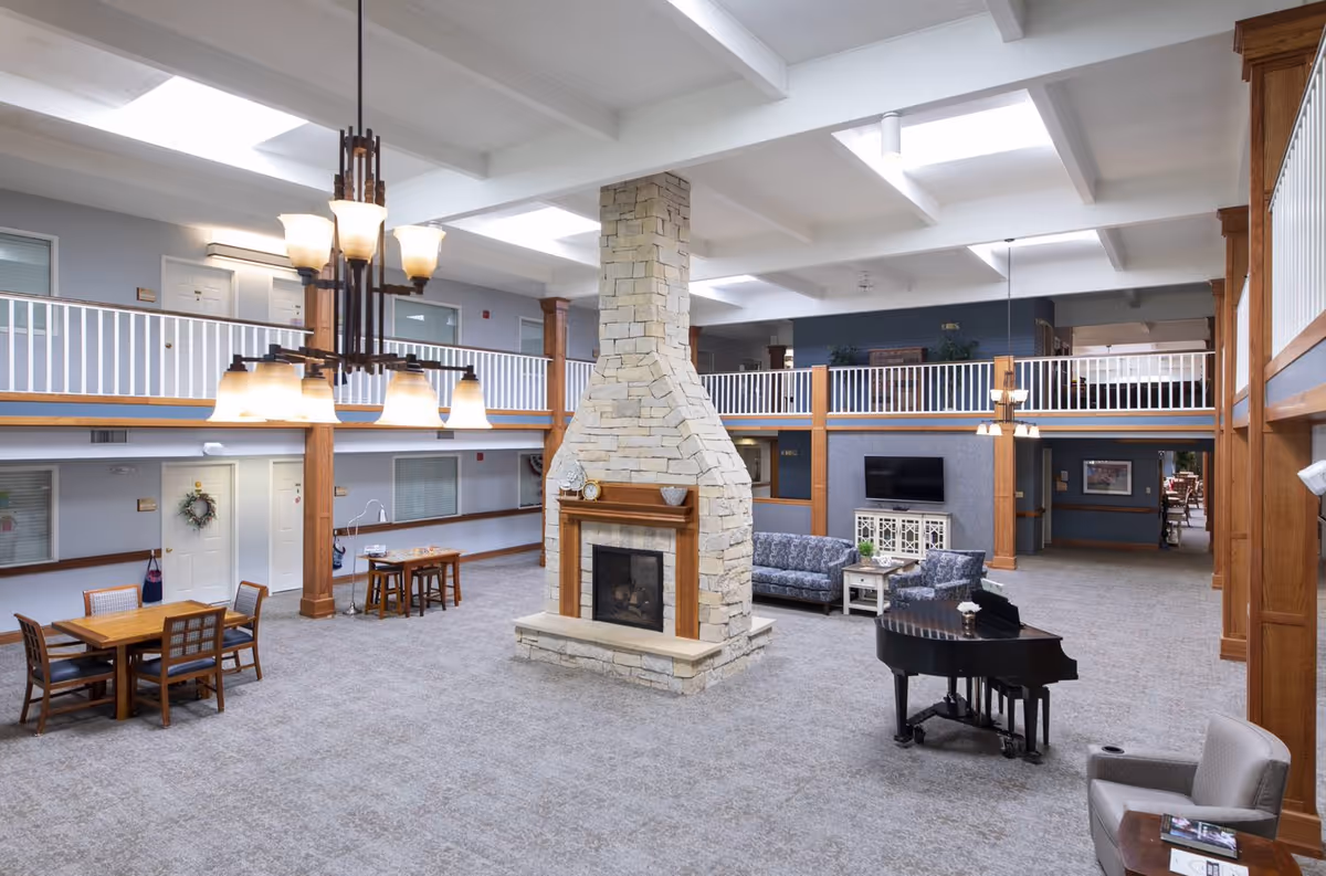 Spacious senior living facility common area with a large stone fireplace in the center, surrounded by seating including sofas, a grand piano, and dining tables. The room has high ceilings with skylights, wooden beams, and a second-floor balcony with white railings overlooking the area.