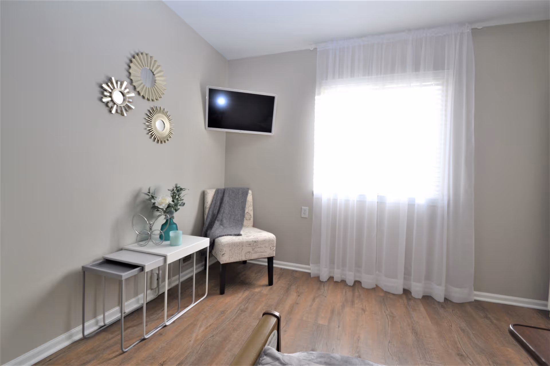 A bright room with light gray walls and wood flooring featuring a window with sheer white curtains. There is a small white nesting table set with decorative items including a vase with flowers and a candle. A beige upholstered chair with a gray throw blanket is placed next to the table. Three decorative round mirrors are mounted on the wall above the table. A flat-screen TV is mounted on the adjacent wall.