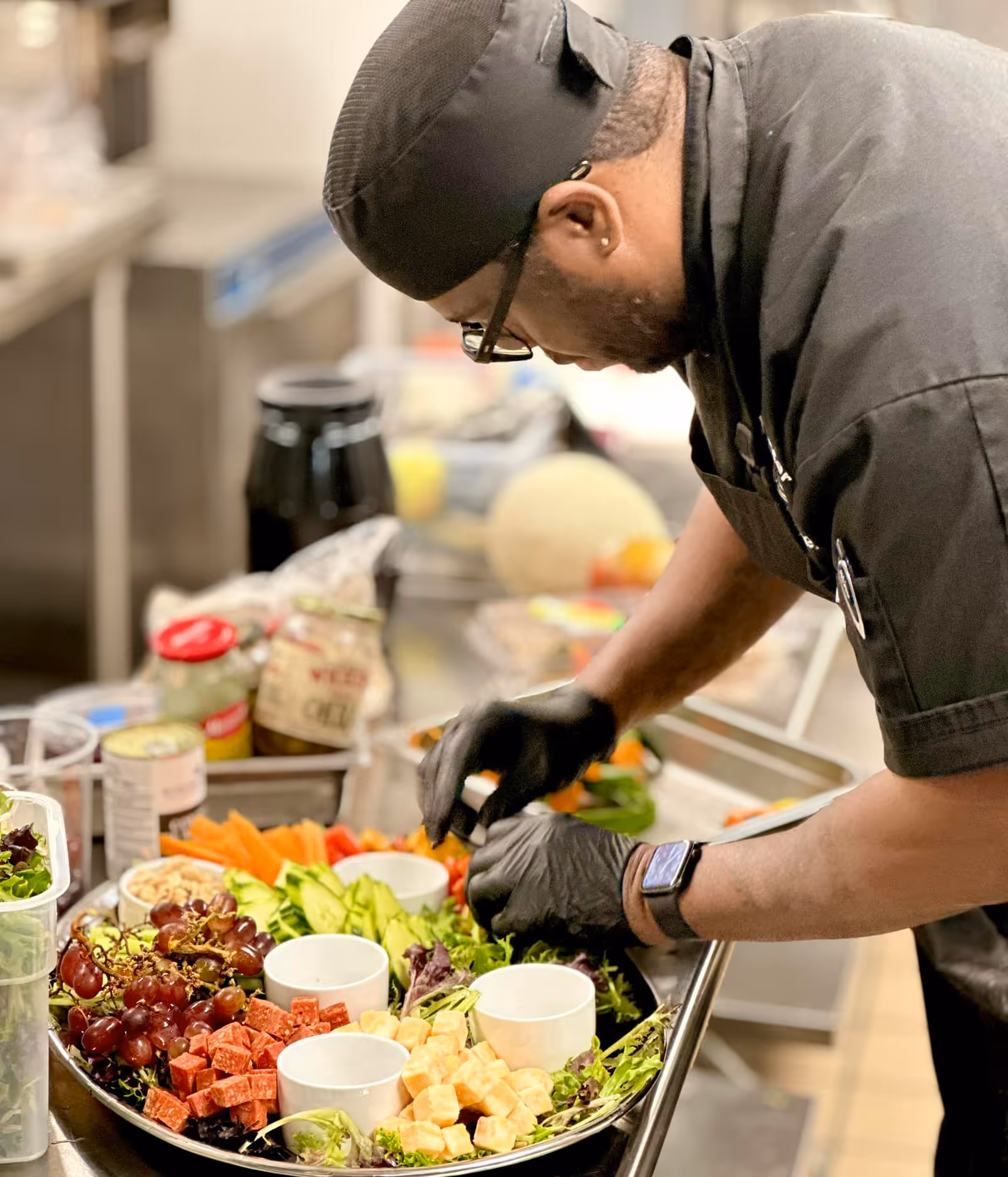 A chef wearing black gloves and a hat arranges a large platter of fruit, cheese, and vegetables in a commercial kitchen.