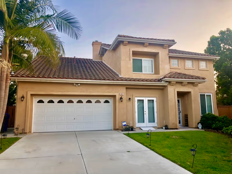 A two-story beige stucco house with a red tile roof, a double garage door, a driveway, and a well-maintained green lawn with small garden lights. There are palm trees and other greenery around the house, and the sky is clear with a soft evening light.