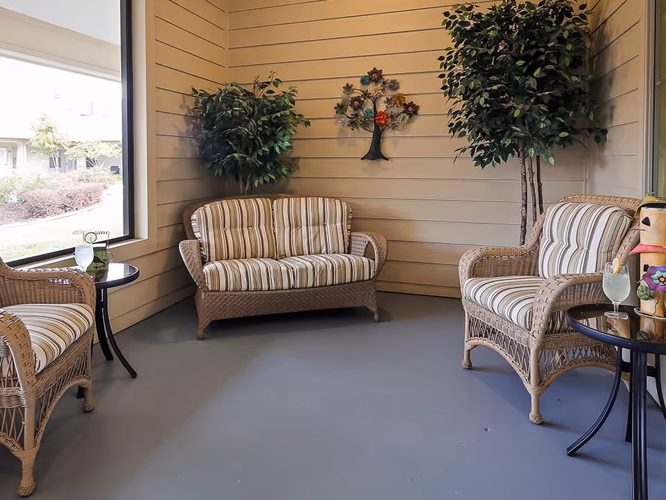 A cozy indoor seating area with wicker furniture including a loveseat and two armchairs with striped cushions. There are two small round black tables, each holding a glass of a light-colored beverage. The walls are beige with horizontal paneling, decorated with a metal tree wall art. Two tall artificial plants are placed in the corners. A large window shows an outdoor view with bushes and a building.