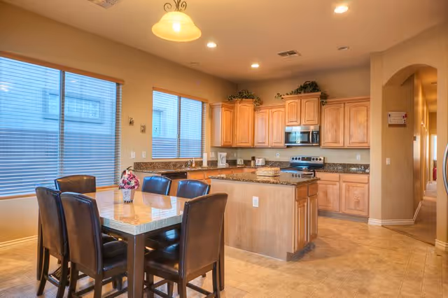 Open kitchen with wooden cabinets, a central island and a dining table with six chairs by large windows.