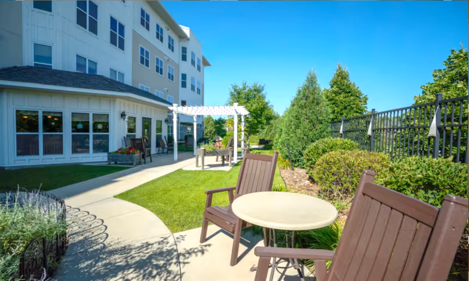 Outdoor patio area at York Gardens with a round table and wooden chairs on a concrete pathway surrounded by green grass, bushes, and trees. The building with multiple windows is visible on the left side under a clear blue sky.