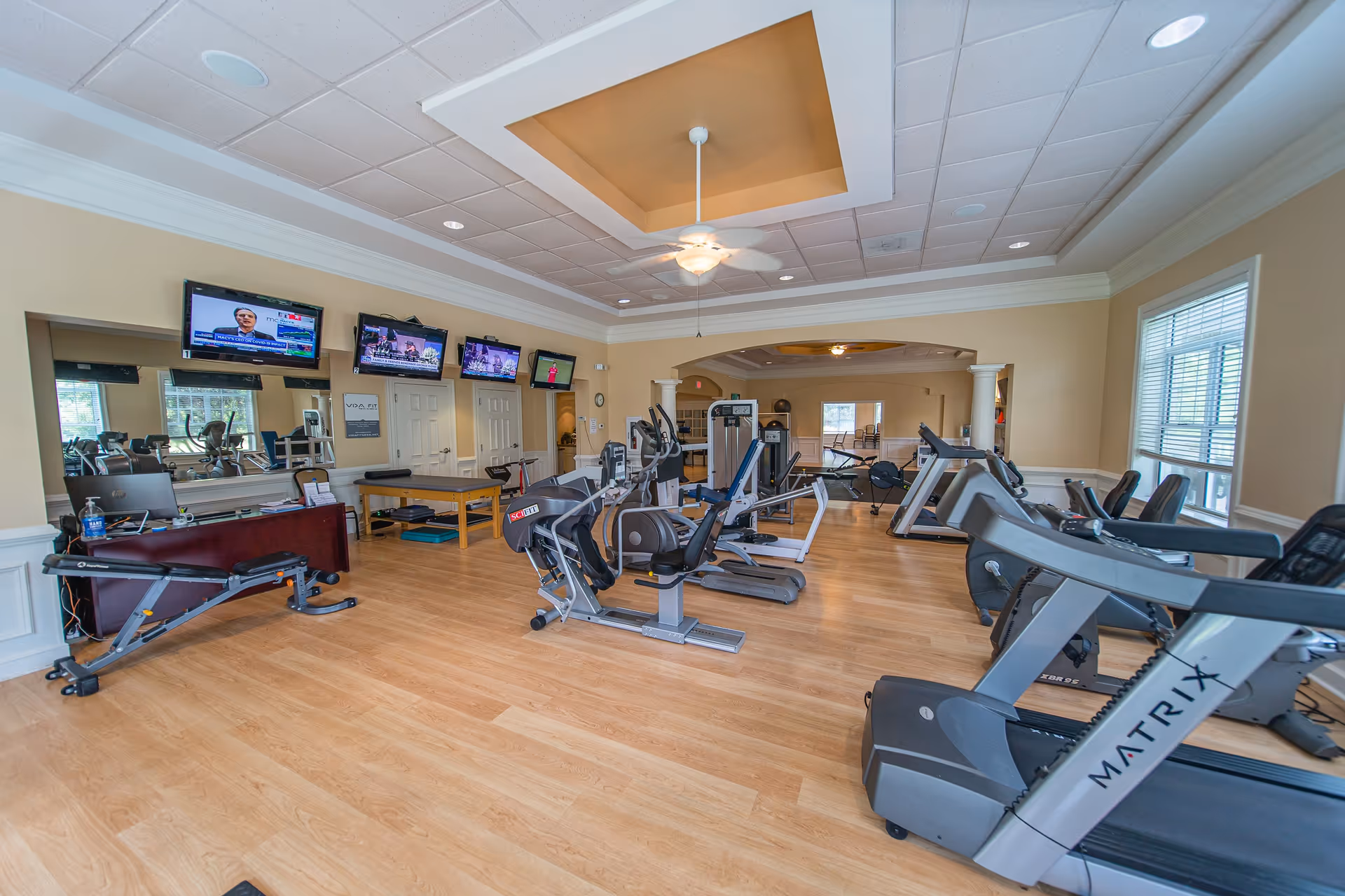A spacious fitness room with various exercise equipment including treadmills, stationary bikes, and weight machines. The room has light wood flooring, beige walls, a ceiling fan, and multiple flat-screen TVs mounted on the wall. Large windows allow natural light to enter the space.