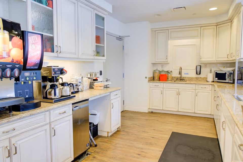 A bright kitchen area with white cabinets, granite countertops, and wooden flooring. The kitchen features a coffee and beverage station with multiple coffee pots, a microwave, a sink, and various kitchen supplies neatly arranged on the counters.