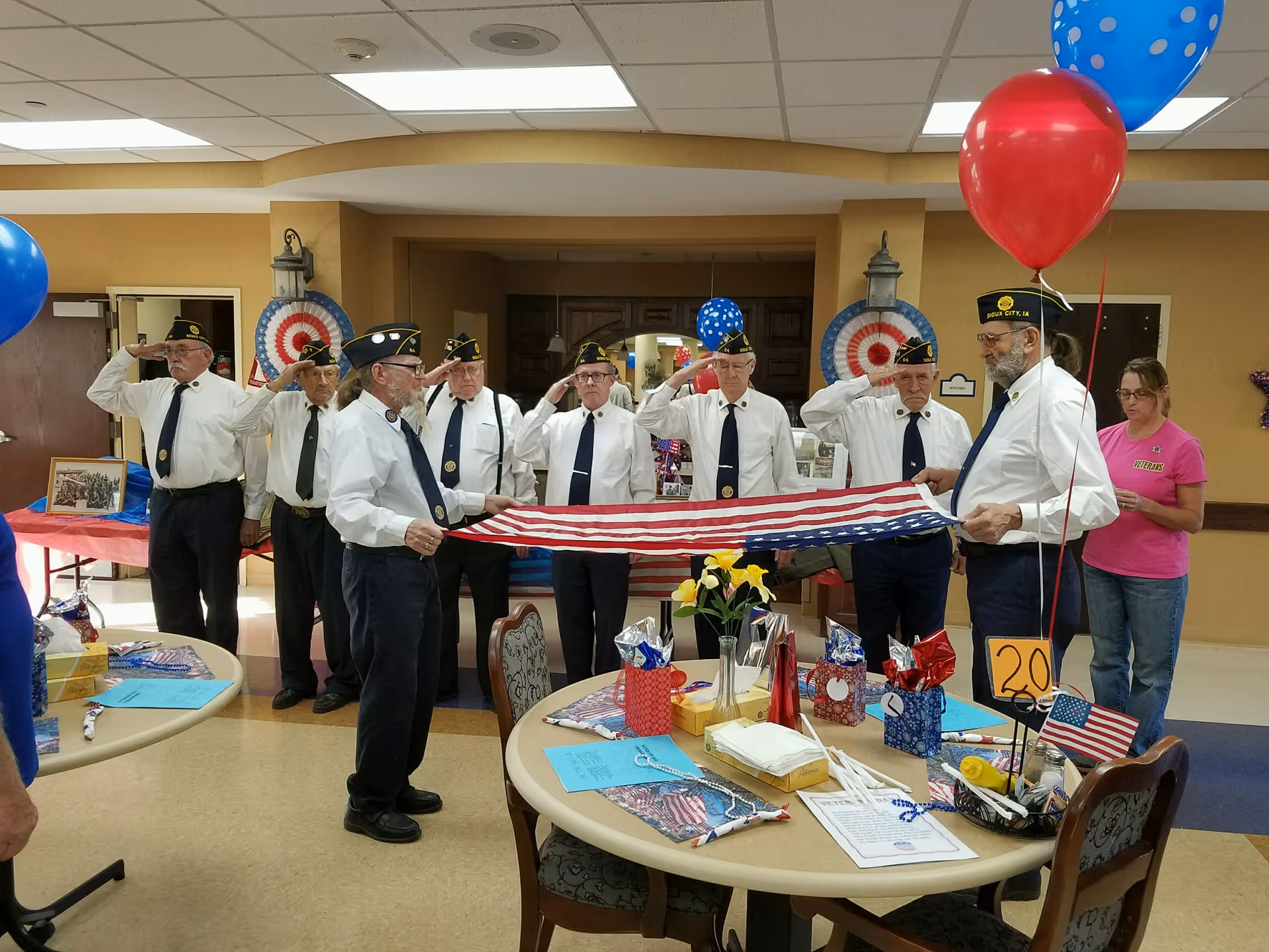 A group of elderly men in white shirts, dark pants, and military hats are standing indoors, saluting while holding a folded American flag. The room is decorated with patriotic items including red, white, and blue balloons, and a table with various decorations and flowers. A woman in a pink shirt stands nearby observing.