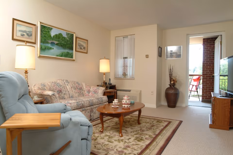 Bright living room with a floral sofa, blue recliner, wooden coffee table, lamps, artwork on the wall, and a doorway leading to a small balcony.