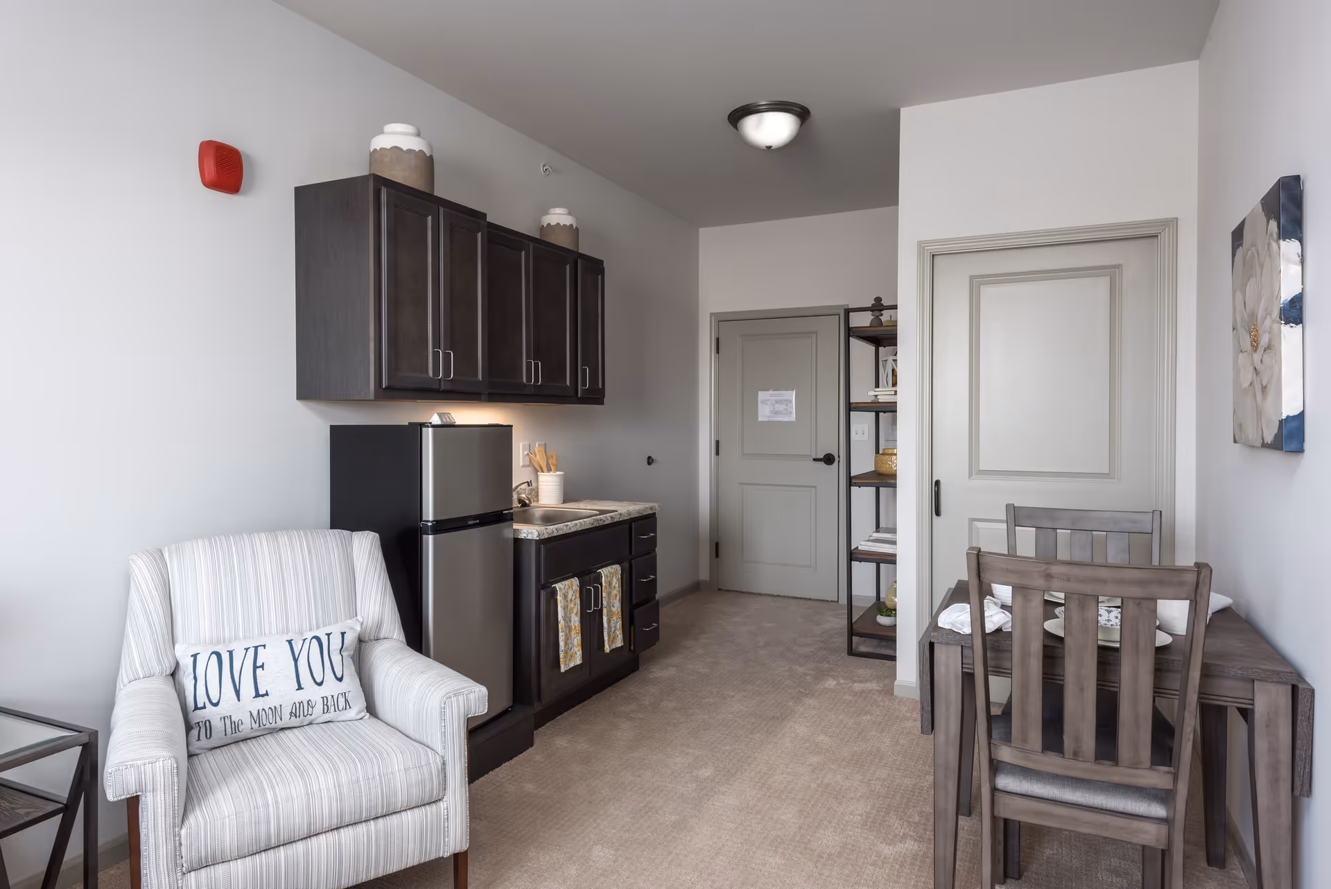 Compact senior living suite interior with a kitchenette, refrigerator, armchair and a small dining table.