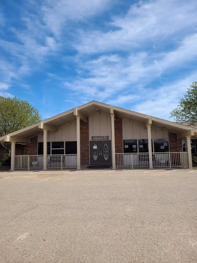 Front exterior view of Enterprise Estates Nursing Center building with a peaked roof, brick and beige siding, double glass doors, and a railing along the front porch. The sky is blue with some clouds and there are trees on either side of the building.