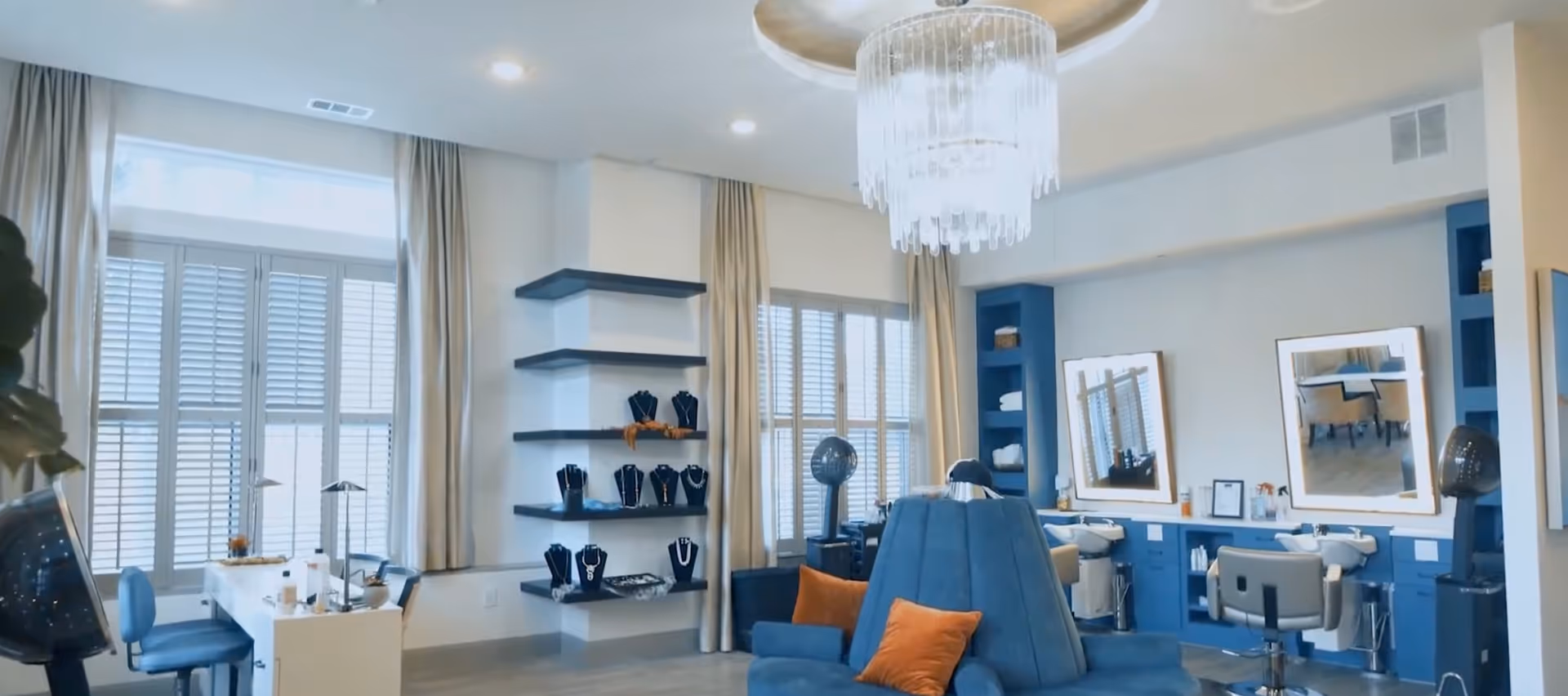 Bright salon-style interior with blue styling chairs and seating, mirrors and workstations, shelving displaying jewelry, and large shuttered windows under a chandelier.