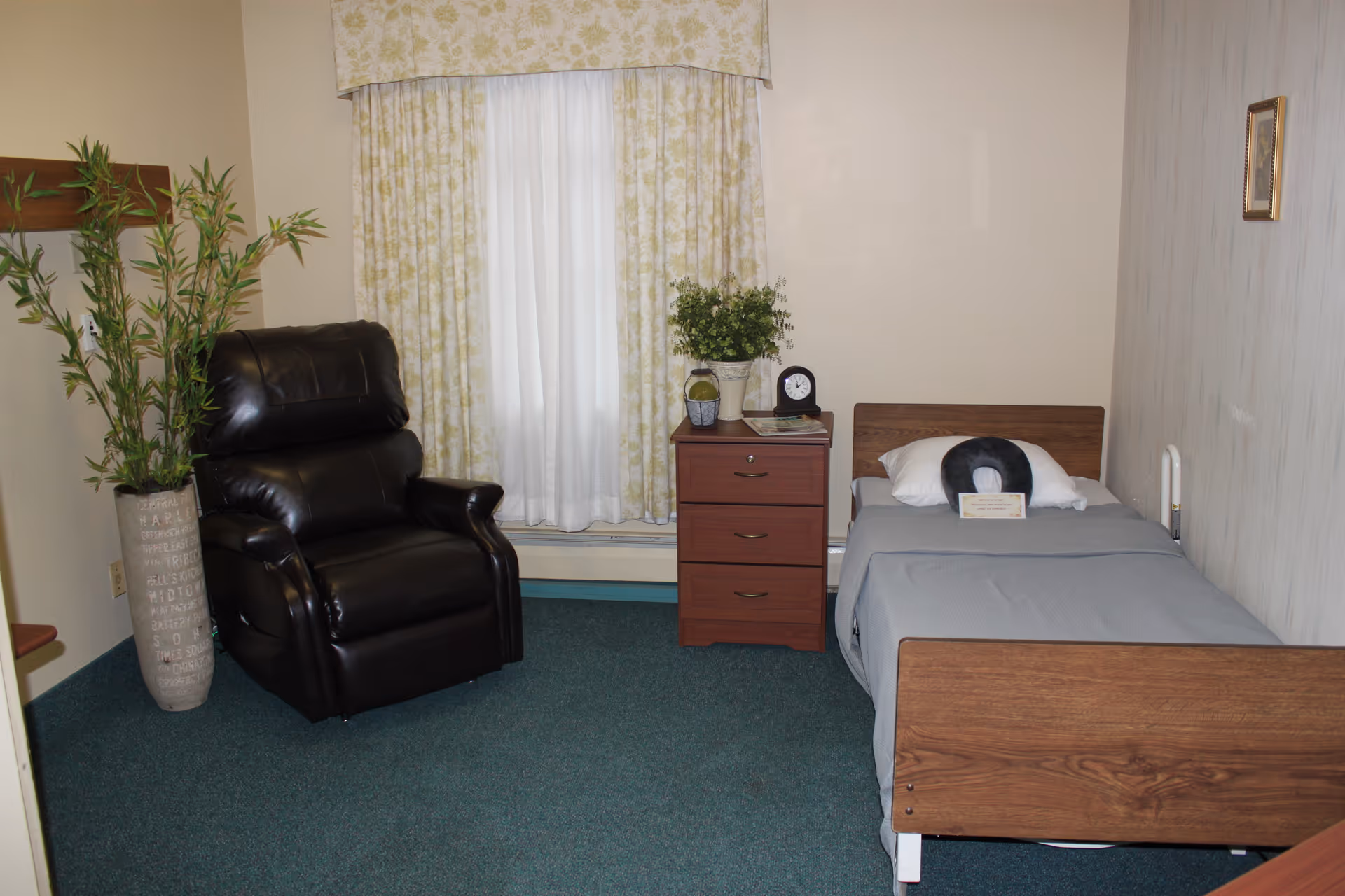 A small, simple bedroom in a care facility featuring a single wooden bed with gray bedding and a pillow with a black neck support cushion. Next to the bed is a wooden nightstand with three drawers, holding a small clock, a potted plant, and some papers. To the left of the nightstand is a black leather recliner chair. A tall potted plant is placed near the chair. The room has green carpet, cream-colored walls, and a window with floral-patterned curtains.