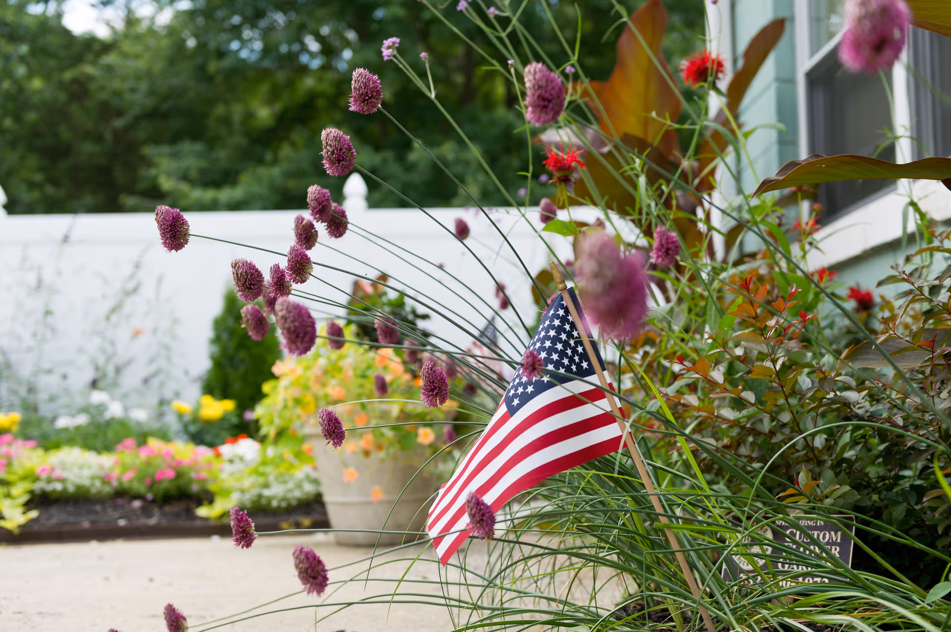 Close-up view of a garden bed with purple flowers and an American flag planted among green foliage, with a blurred background of more colorful flowers and a white fence.