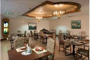 A dining room with multiple tables and chairs arranged neatly. Each table is set with placemats, napkins, and small flower vases. The room has a decorative ceiling with a chandelier and framed paintings on the walls.