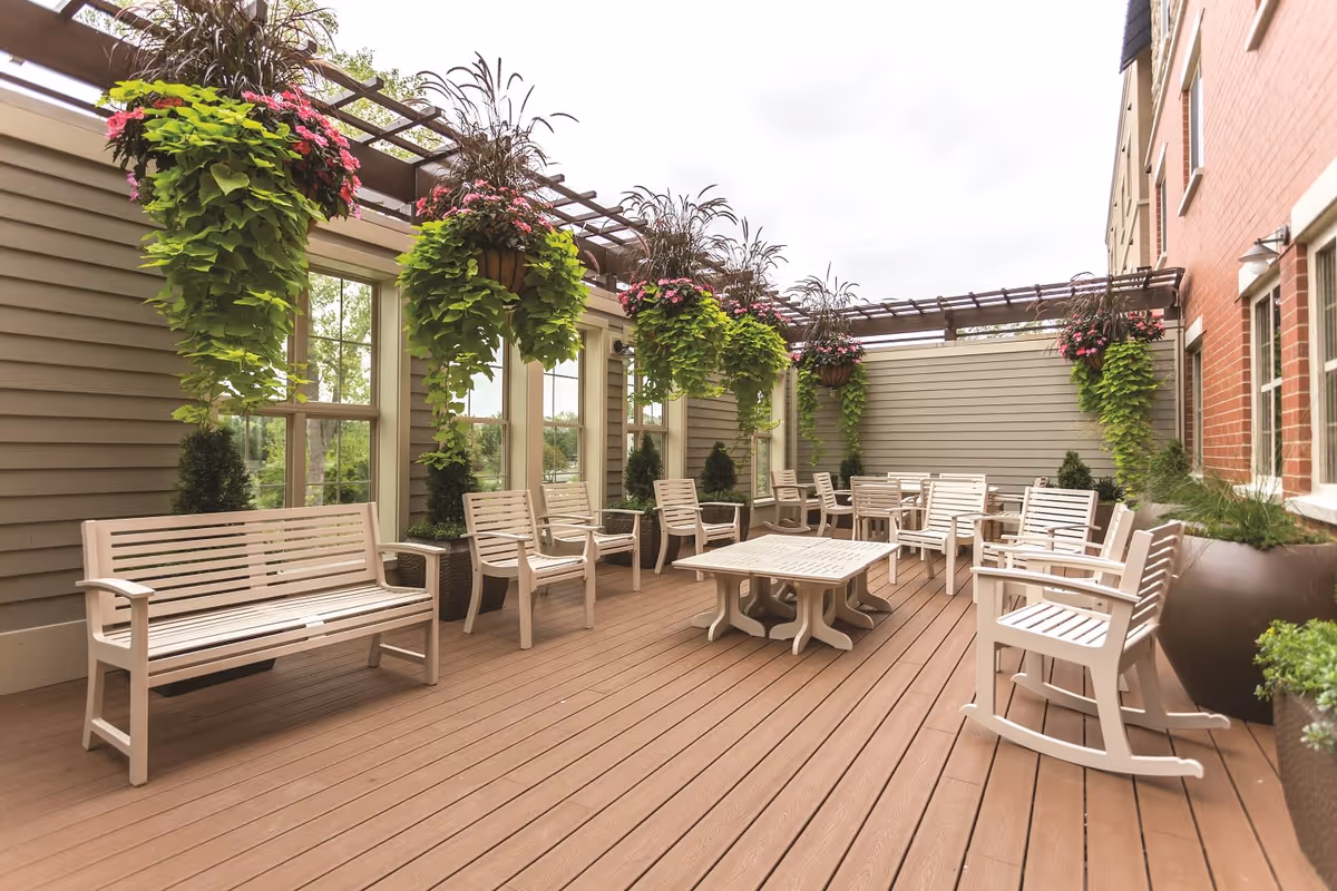 Outdoor patio area with multiple beige benches, chairs, and a table on a wooden deck. Hanging flower baskets with green foliage and pink flowers are suspended from a pergola above. The patio is enclosed by beige siding walls and a brick building wall with windows.