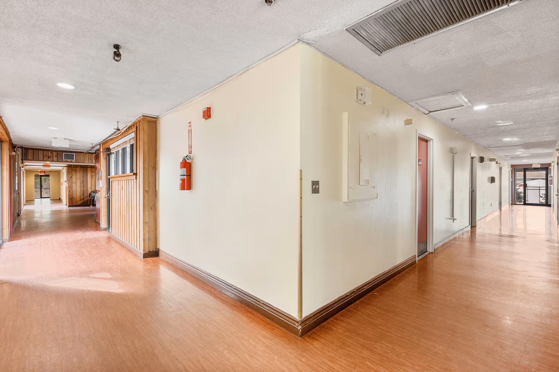 A bright hallway in an assisted living facility with light-colored walls and wood-patterned flooring. The hallway has multiple doors on the right side and a fire extinguisher mounted on the wall. The ceiling has recessed lighting and ventilation panels. At the end of the hallway, there are glass doors allowing natural light to enter.