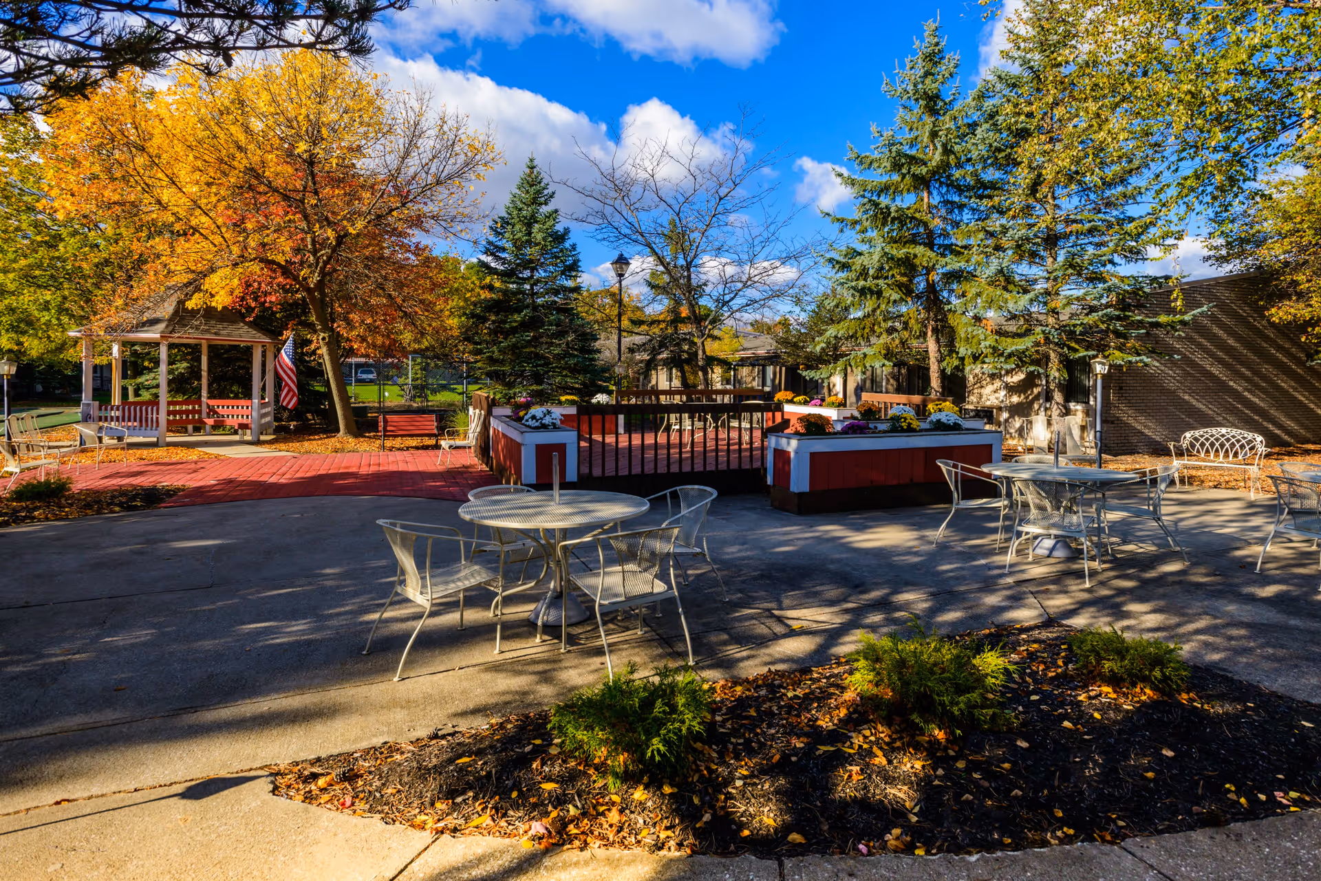 Outdoor patio area at Woodside Senior Living with metal tables and chairs on a concrete surface, surrounded by trees with autumn foliage, a gazebo with an American flag, and flower planters. The sky is blue with some clouds.