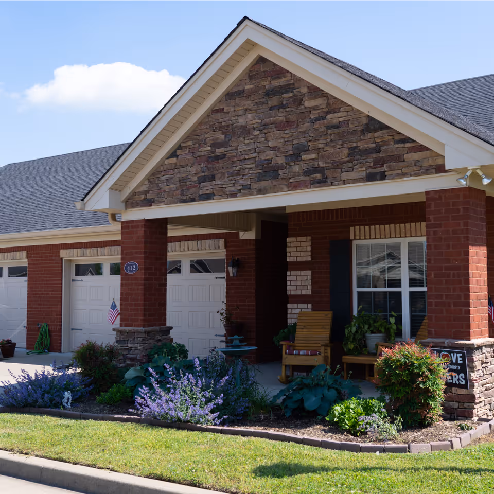 Front entrance and garage of a brick senior-living building with a stone gable, flowering beds, and wooden chairs on the covered porch.