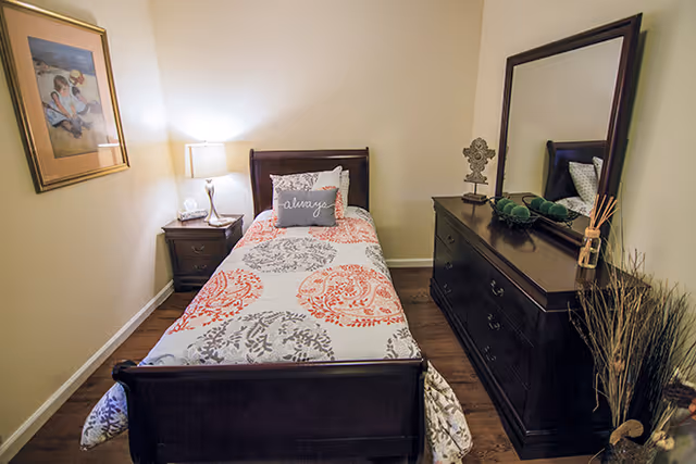 Small furnished bedroom with a twin bed, nightstand and lamp on the left, and a dresser with mirror on the right.