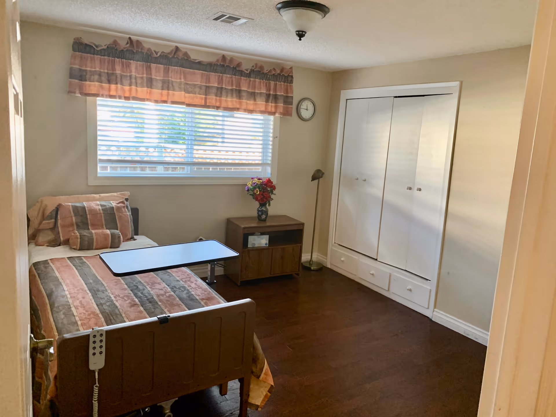 A small bedroom with a single bed featuring a striped bedspread and pillows. There is a movable overbed table on the bed. A window with blinds and a valance is on the wall behind the bed. Next to the bed is a wooden nightstand with a vase of flowers and a floor lamp beside it. A wall clock is mounted above the nightstand. Across from the bed is a white closet with double doors and drawers underneath. The room has wooden flooring and beige walls.