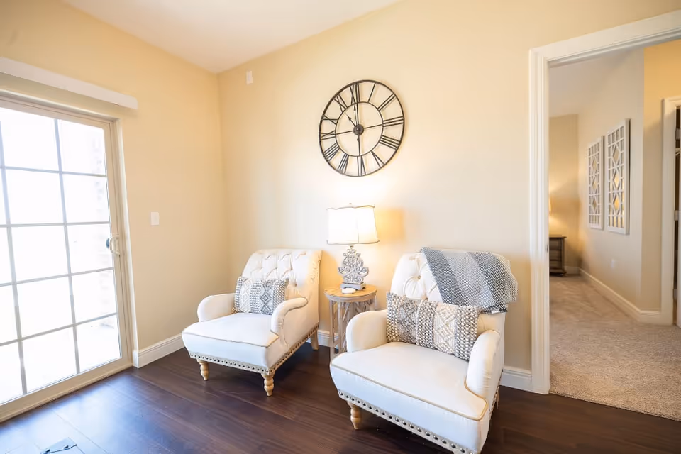 A cozy sitting area in a senior living facility with two cream-colored armchairs adorned with patterned pillows and a throw blanket. Between the chairs is a small round wooden side table with a decorative lamp. A large wall clock with Roman numerals hangs above the chairs. To the left, there is a sliding glass door letting in natural light, and to the right, an open doorway leads to another room with carpeted flooring and wall decorations.