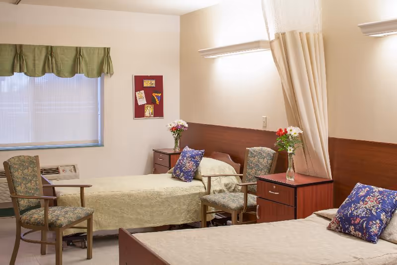 A shared bedroom in a senior living facility with two beds, each with a floral pillow and light-colored bedding. Between the beds are two wooden nightstands with vases of flowers. Two upholstered chairs are positioned near the beds. A window with a green valance and a bulletin board with pinned notes are visible on the wall.