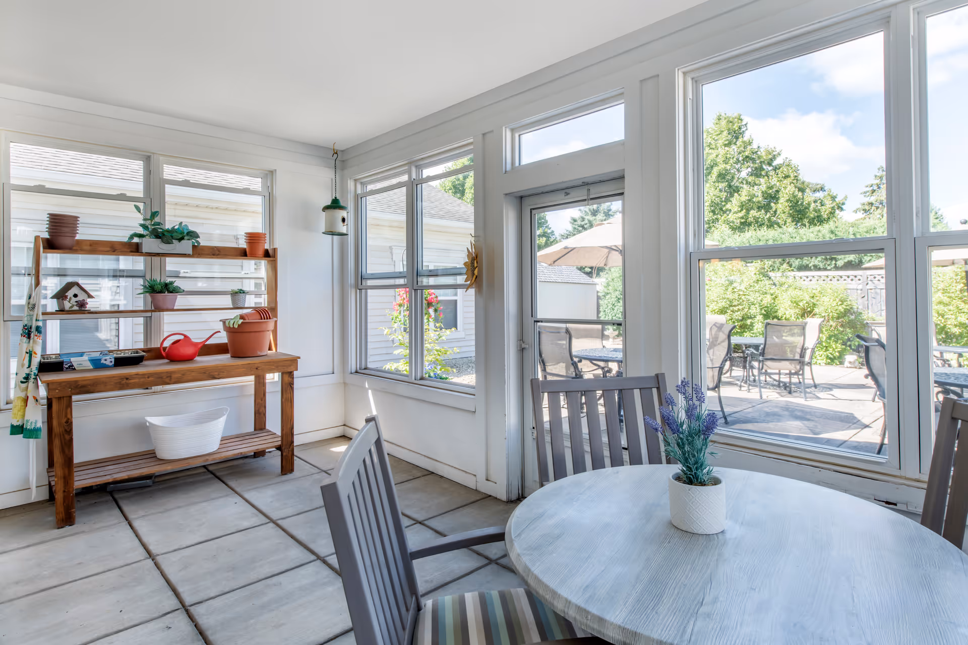 A bright sunroom with large windows and a glass door leading to an outdoor patio area. Inside, there is a round wooden table with a small potted plant and two chairs with striped cushions. Against one wall is a wooden shelf holding various small potted plants, a watering can, and gardening items. Outside, patio tables and chairs with umbrellas are visible, surrounded by greenery and trees.