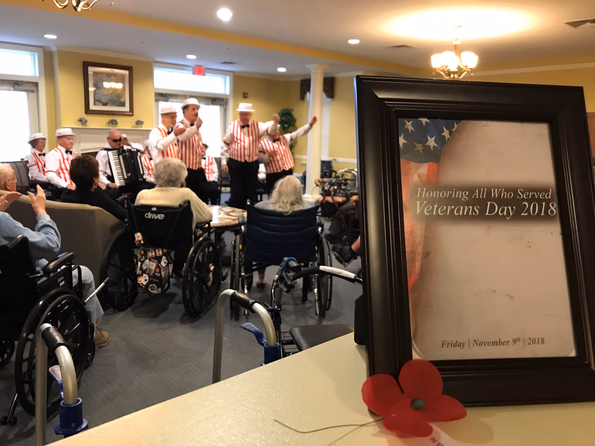A group of elderly residents, some in wheelchairs, watch a musical performance by men dressed in red and white striped vests and white hats in a common room with yellow walls and large windows. In the foreground, a framed sign reads 'Honoring All Who Served Veterans Day 2018 Friday | November 9th | 2018' with a red poppy flower placed in front of it.