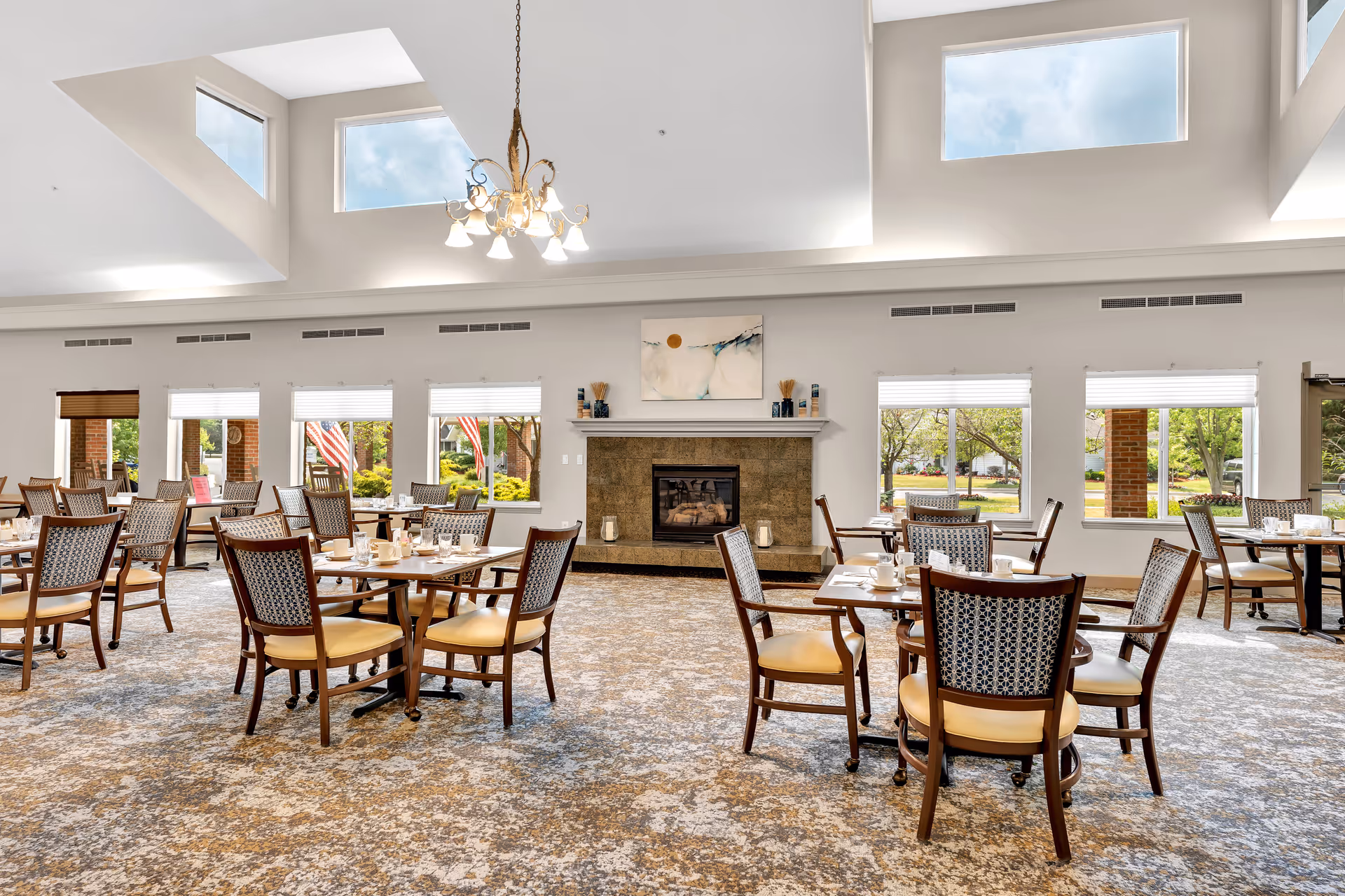 A spacious dining room with multiple wooden tables and chairs arranged neatly. The room features large windows letting in natural light, a fireplace centered on the far wall with decorative items on the mantel, and a chandelier hanging from the ceiling. The carpet has a patterned design, and the walls are painted white.