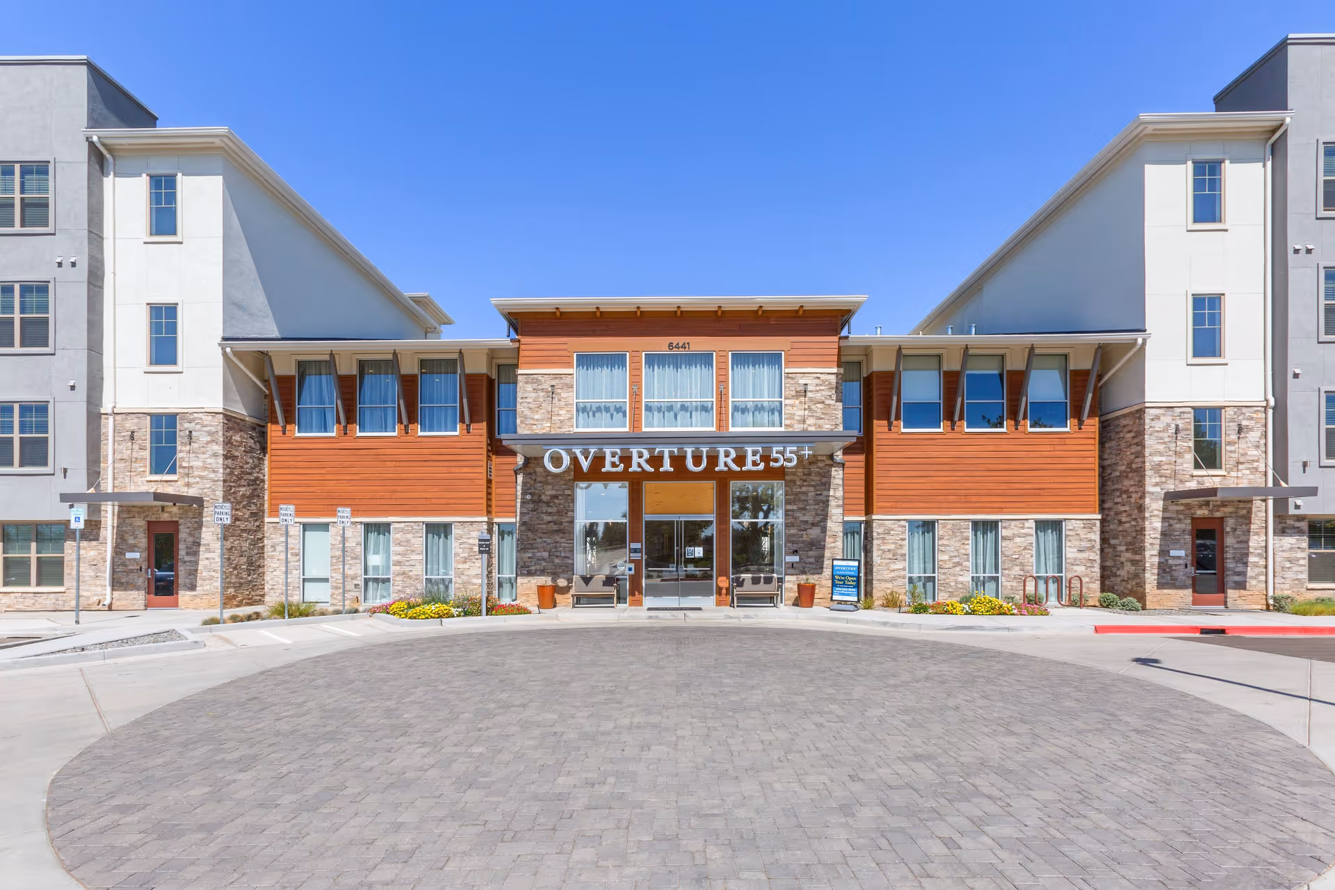 Front exterior view of Overture Albuquerque senior living facility with a clear blue sky, featuring a modern building with stone and wood panel accents, large windows, and a circular driveway in front.