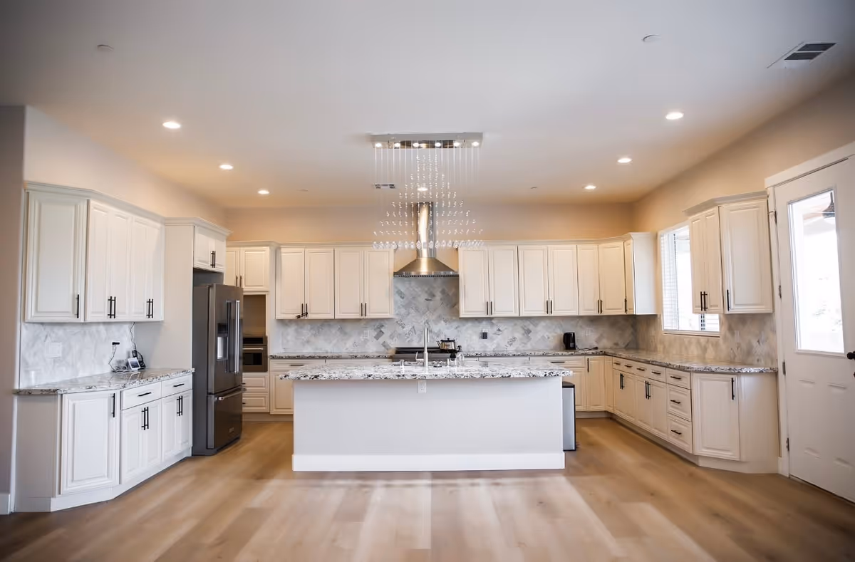 A spacious, modern kitchen with white cabinetry, a large central island with a granite countertop, stainless steel refrigerator, built-in oven, and a stainless steel range hood. The kitchen has light wood flooring, recessed lighting, and a decorative chandelier above the island. There is a door and window on the right side allowing natural light in.