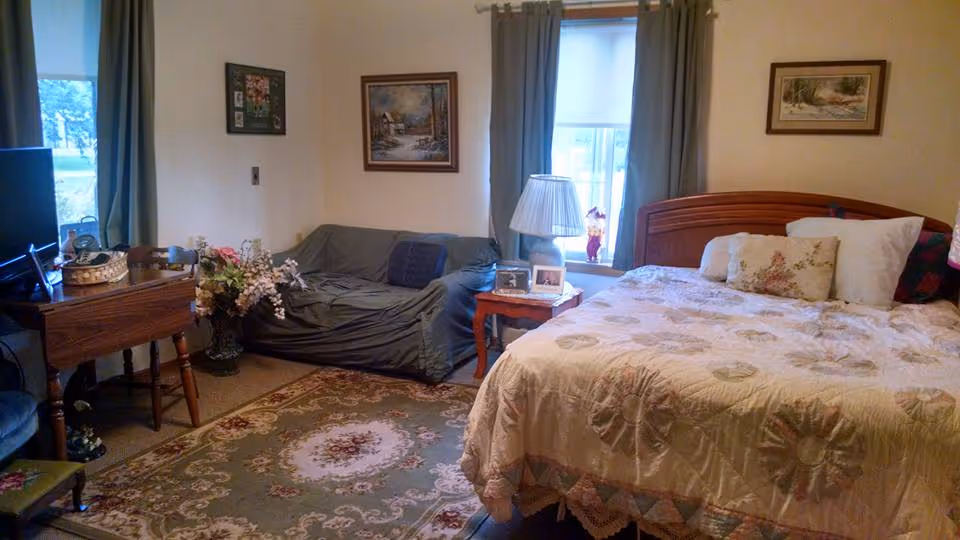 A cozy bedroom with a large bed covered in a floral quilt, a small wooden side table with a lamp and framed photos, a gray couch against the wall, a wooden table with chairs, floral carpet, and framed paintings on the walls. Two windows with curtains allow natural light into the room.