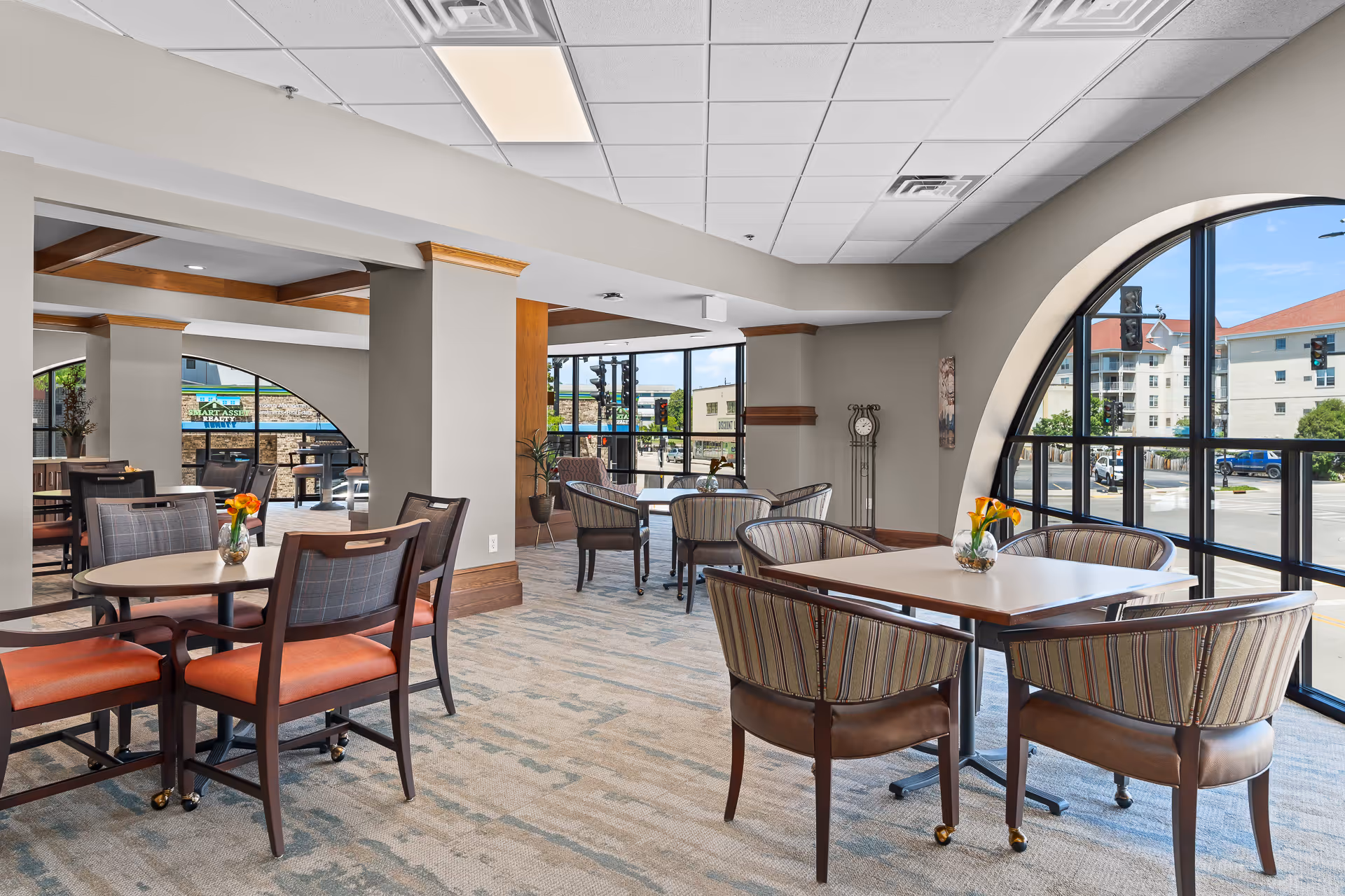 A bright and spacious dining area with multiple tables and chairs arranged neatly. The room features large arched windows allowing natural light to fill the space and providing a view of the street outside. Each table has a small vase with flowers, and the interior has a modern design with neutral tones and wooden accents.