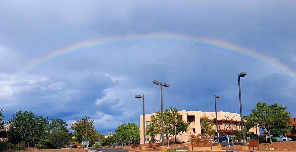 A wide outdoor view of a building with southwestern architectural style under a cloudy sky with a vibrant rainbow arching across the sky. The scene includes several trees, street lamps, parked cars, and benches in the foreground.