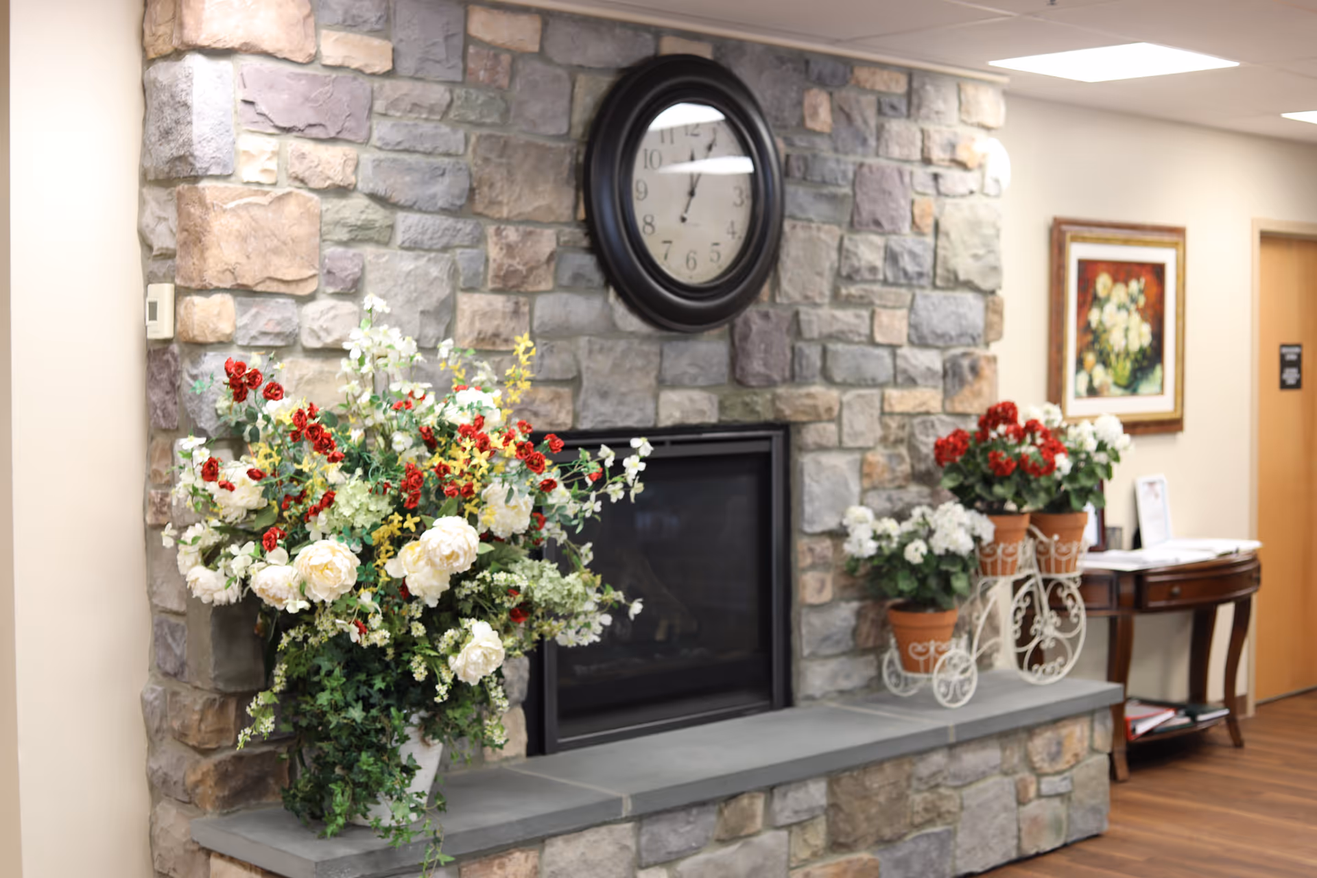 Interior view of a stone fireplace with a black round clock mounted above it. The fireplace mantel is decorated with a large floral arrangement featuring white, red, and yellow flowers on the left side, and three potted plants with red and white flowers on a decorative white metal stand on the right side. In the background, there is a wooden table with papers and a framed floral painting on the wall. The floor is wooden and the ceiling has recessed lighting.