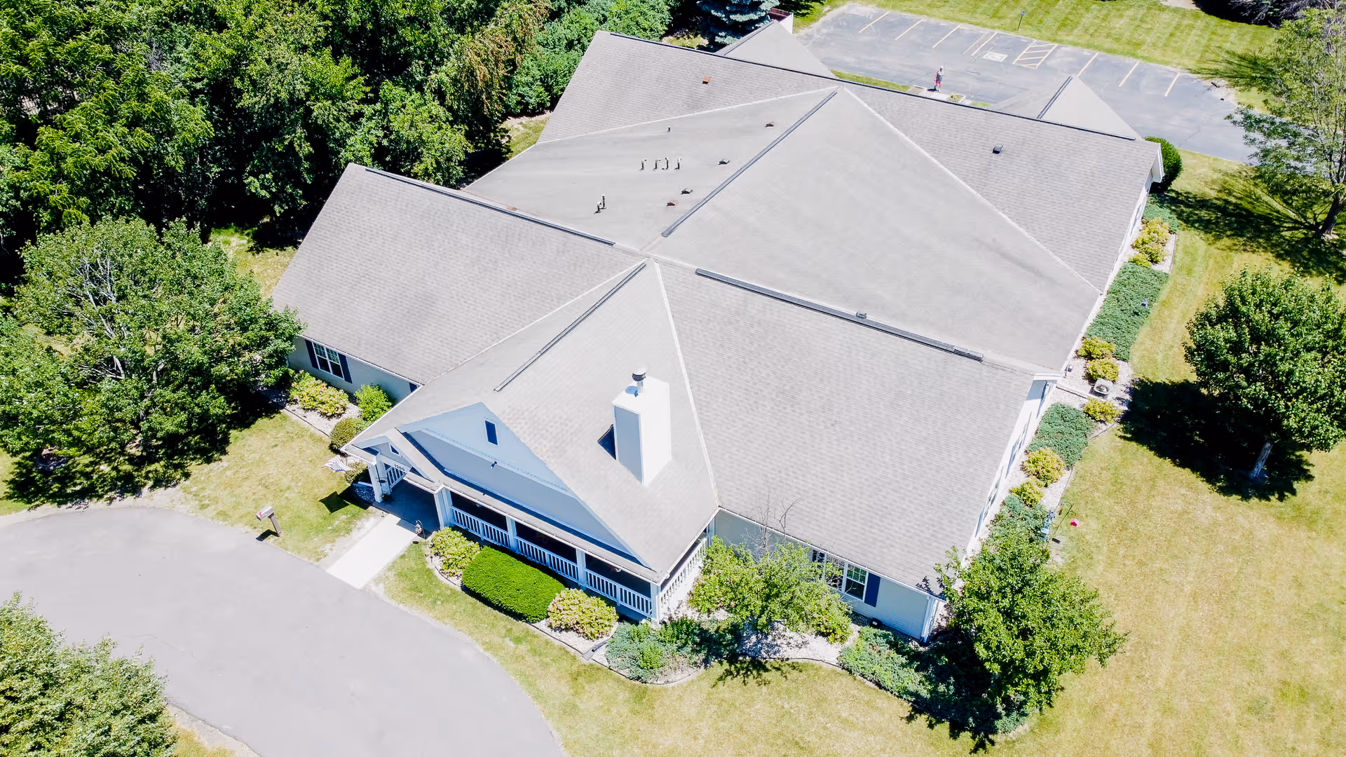 Aerial view of a single-story assisted living building with a gabled roof, front porch, surrounding lawns, trees, and a parking area.