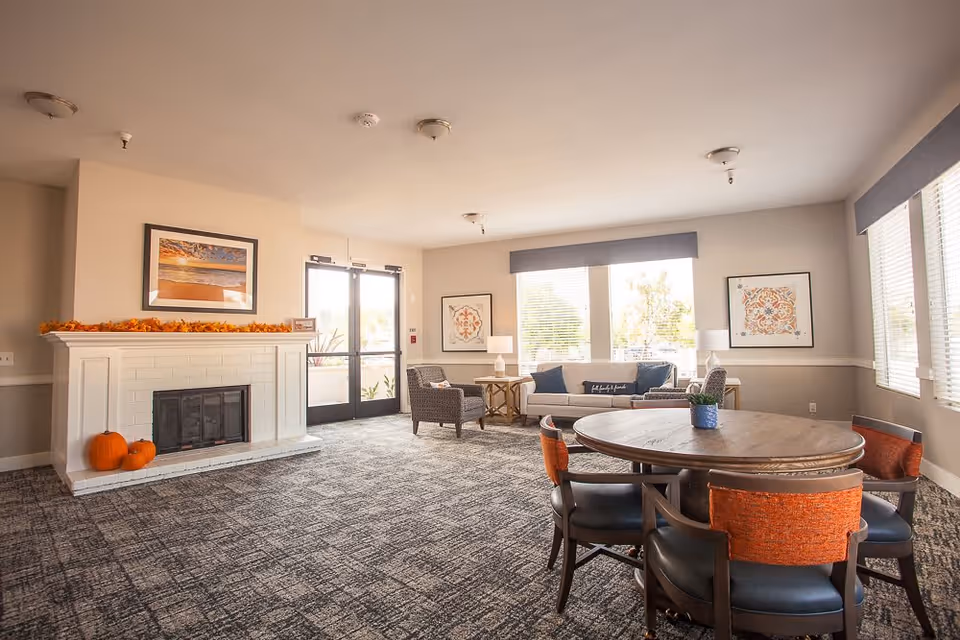 Bright communal living room with a fireplace, seating area, and round table by large windows.