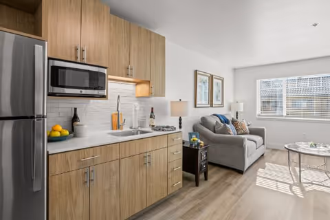 A modern studio apartment interior featuring a compact kitchen with wooden cabinets, a stainless steel refrigerator, microwave, and sink. Adjacent to the kitchen is a cozy living area with a gray sofa, side table with a lamp, framed artwork on the wall, and a window letting in natural light.