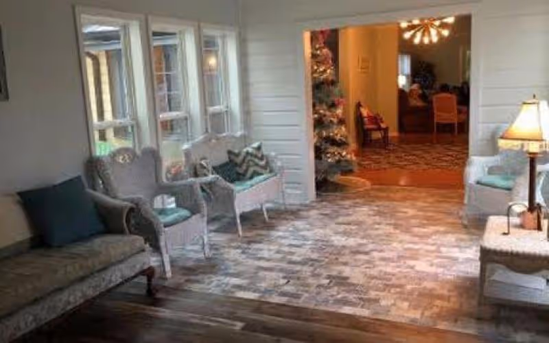 Sunny seating area with wicker chairs, a sofa and a lamp opening into a decorated common room with a Christmas tree.